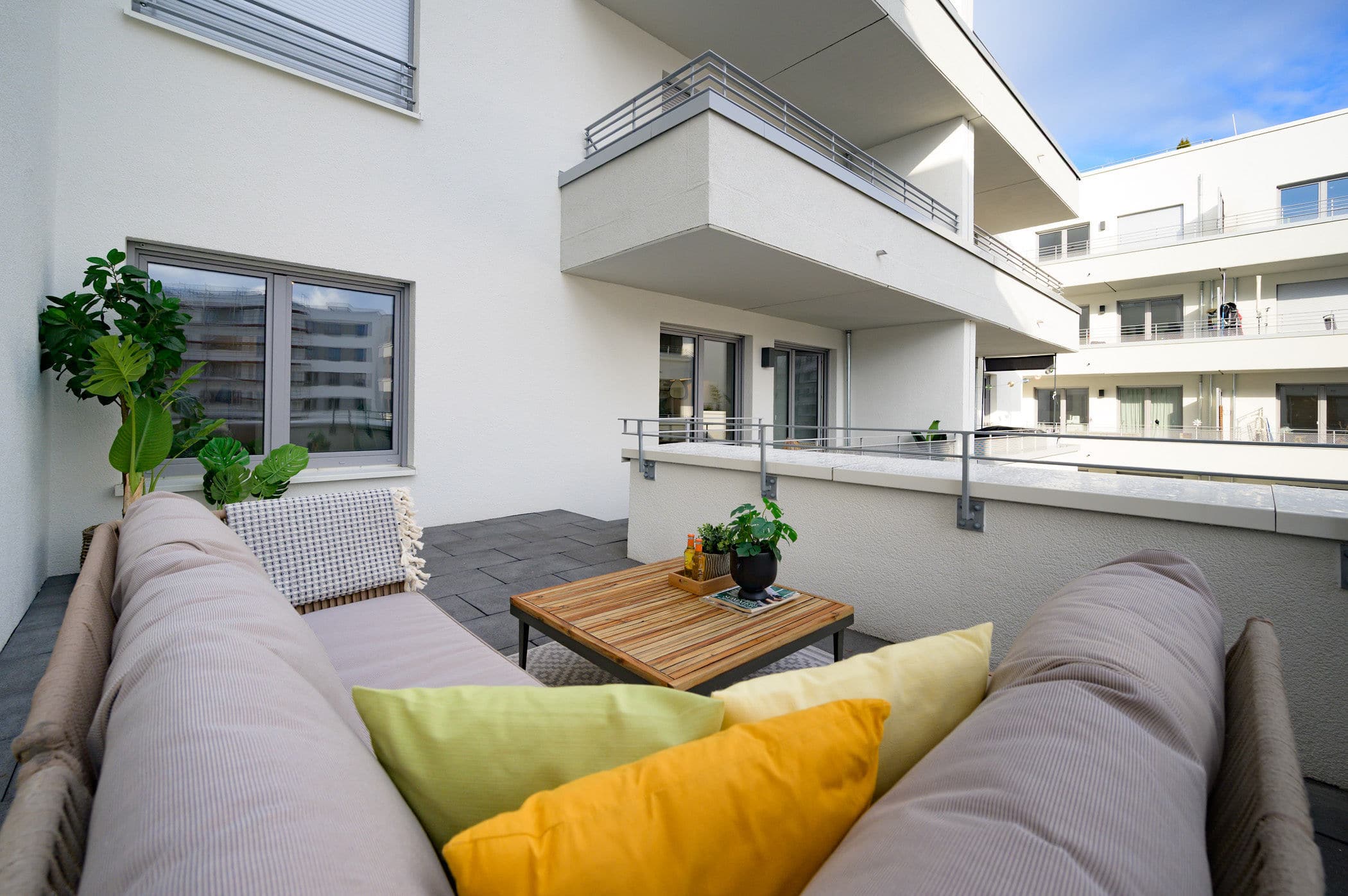 Großzügige Terrasse einer modernen Neubauwohnung mit Lounge-Möbeln, hochwertigem Bodenbelag, Pflanzenelementen und Blick auf die gepflegte Wohnanlage im Innenhof.